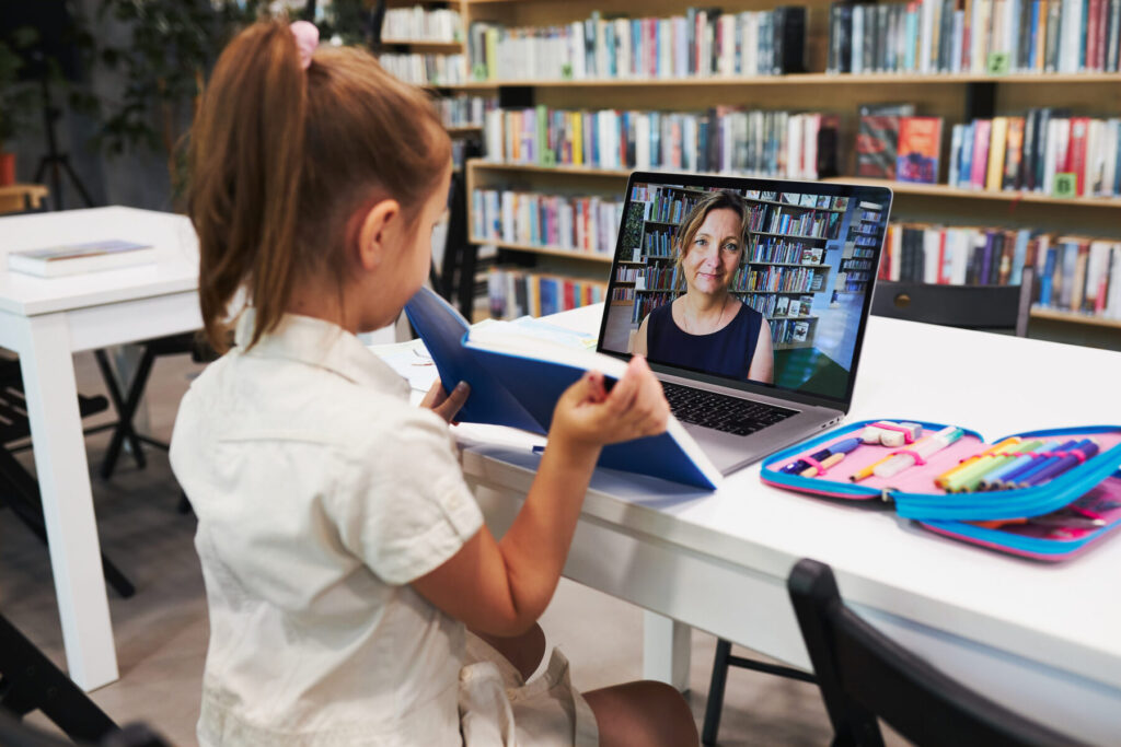 Student little girl having video class remotely with her teacher on laptop sitting at desk