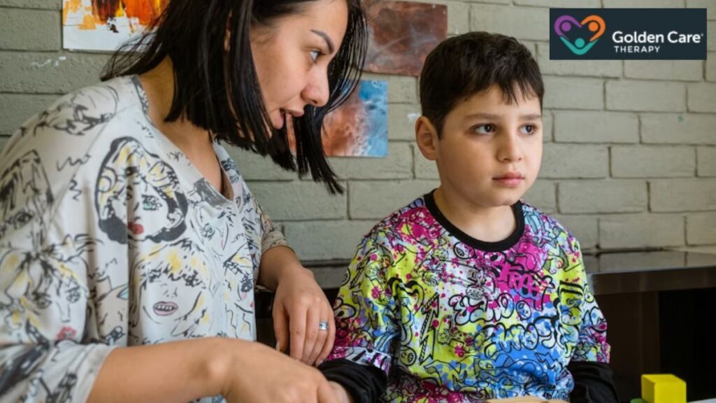 A female therapist sits on a table, her profile facing the camera, talking to a young child. The child stares straight ahead, focused on something in front of him, illustrating a moment of autism regression therapy.