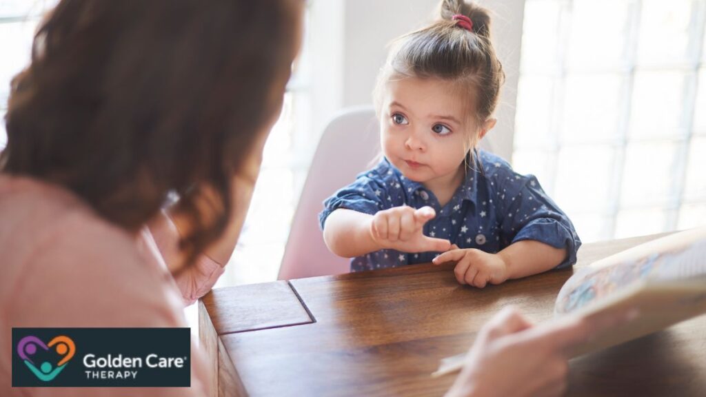 A young girl with autism uses hand motions to communicate with a parent or therapist in a brightly lit dining room.