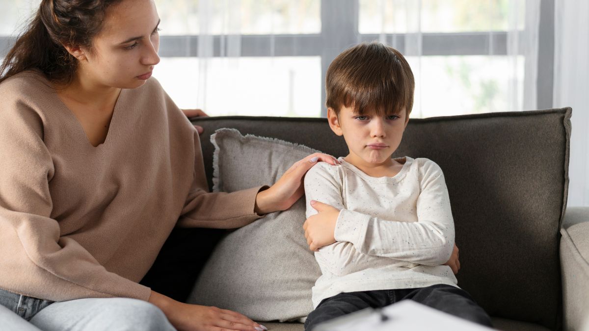 A boy sits on the sofa, his arms tightly crossed in a rigid act of defiance and stubbornness, resisting the gentle coaxing of his caregiver.