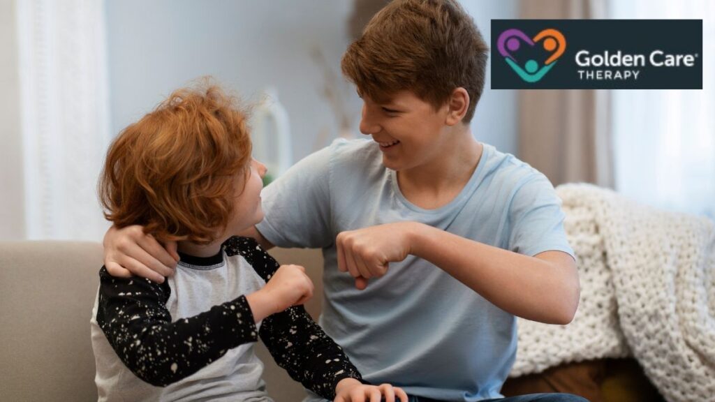 Two boys sit on a sofa, one younger and one older, pressing their knuckles together in a cooperative and joyful moment, illustrating positive interaction amid autism rigidity examples.