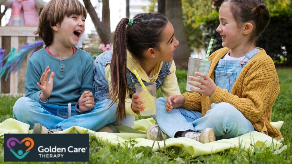 A group of children sit on the grass drinking lemonade, enthusiastically engaging in conversation, illustrating social interaction among children with tone deaf autism.