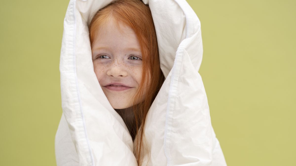 A young girl smiles while looking away from the camera, wrapped in a heavy weighted blanket, with a bright yellow wall in the background.