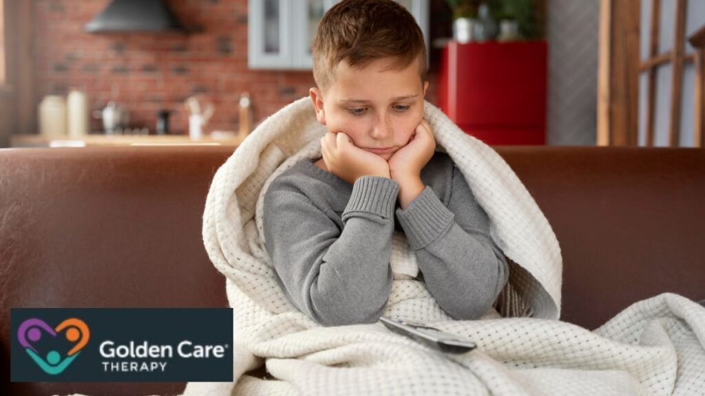 A boy covered by a heavy weighted blanket rests his face in his palms, looking downward and thoughtful, enjoying the calming comfort.