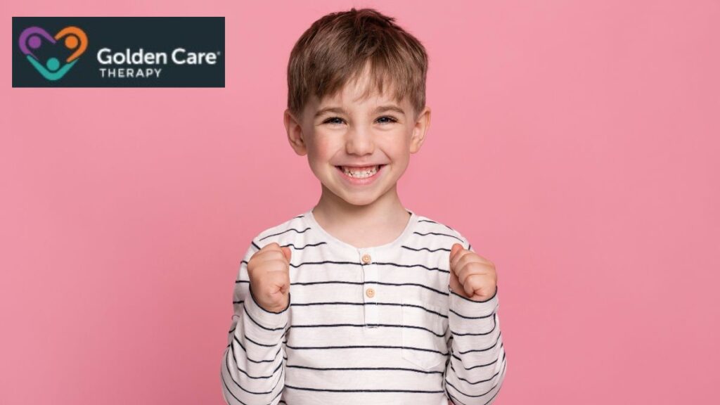 A young autistic boy smiles brightly at the camera against a pink background, his hands raised in small folded fists as he expresses pure joy.