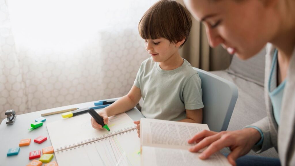 A young boy with autism sits at a table covered with puzzle pieces, preparing to take notes in his notebook while his instructor gently guides him nearby during a behavior modification session.