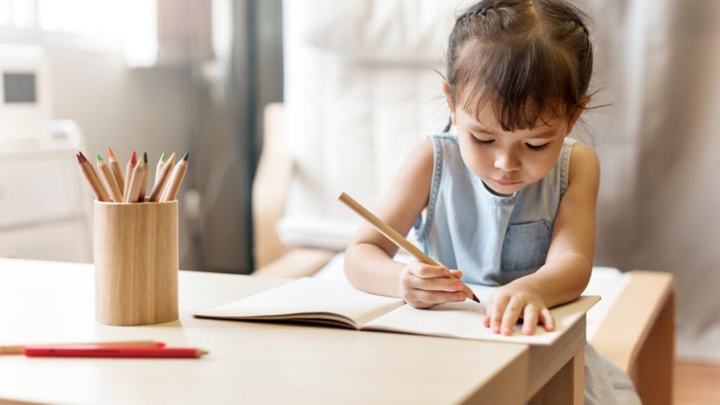 A young girl works on a drawing task at a table, with colored pencils in a cylinder holder beside her, following a daily visual schedule.