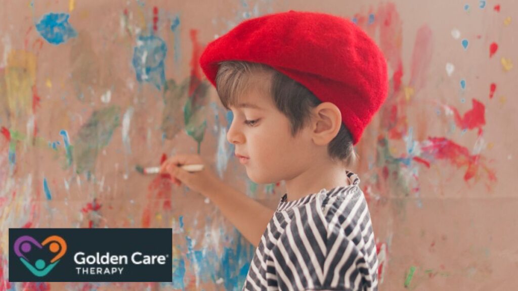 A young boy wearing a red hat paints an abstract image on a propped canvas, looking focused and serious as he works.