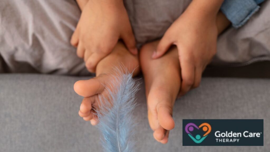 A toddler sits on a carpeted floor while a parent holds them steady. A feather rests against the child’s feet as a soothing sensory alternative to rubbing their feet together.
