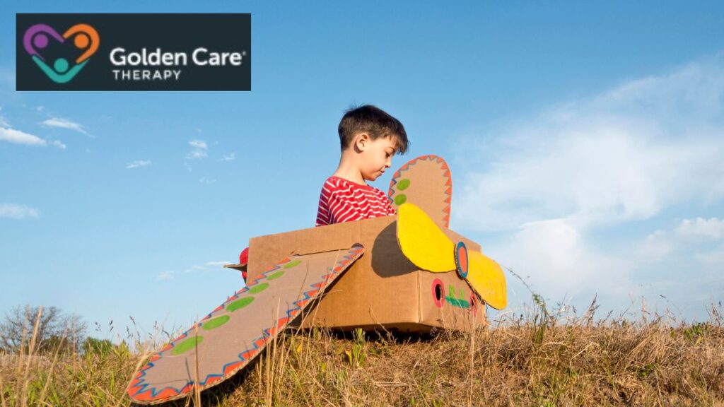 A young boy sits in a makeshift plane made from brightly painted cardboard boxes, playing in the middle of a sunlit farm.
