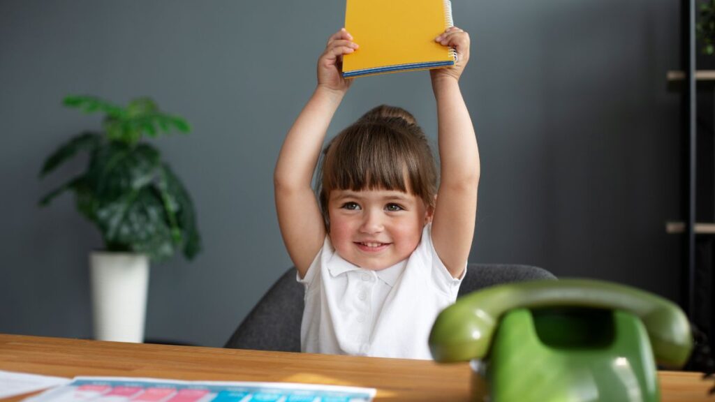 A young girl holds a notepad above her head, appearing busy, sitting at a table with school materials and a green vintage phone, following her daily visual schedule.