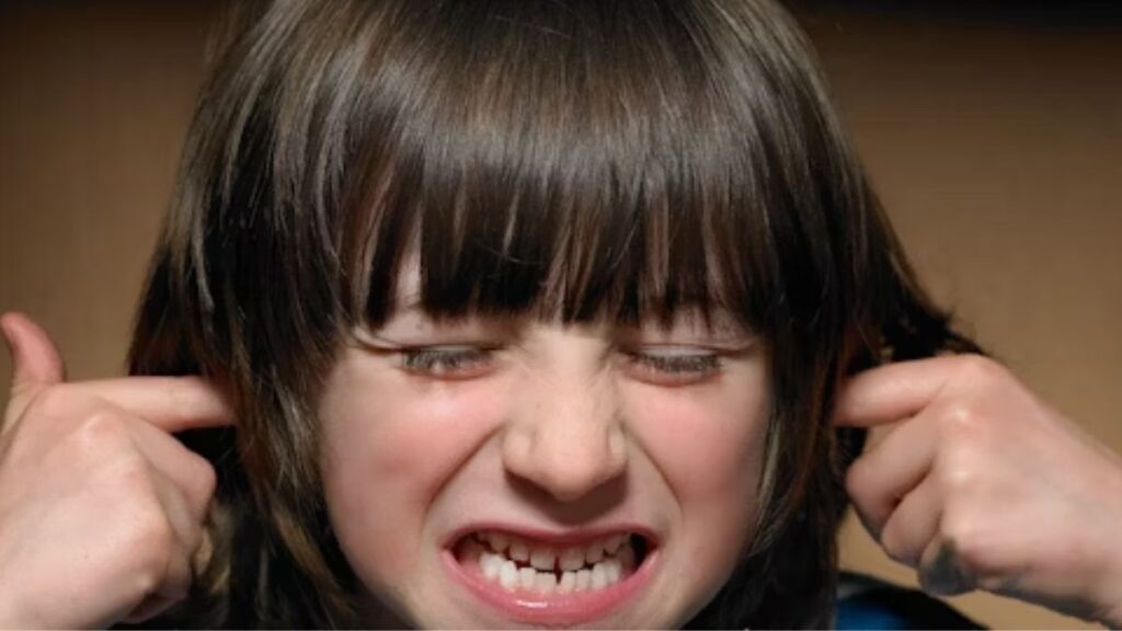 A young boy covers his ears and shows a look of extreme discomfort, struggling with sensory overstimulation.