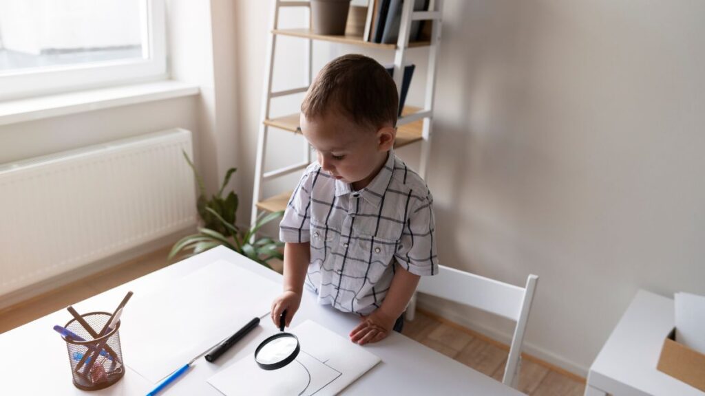 A young boy holds a magnifying glass while working on a project at a table covered with large sheets of paper featuring geometry graphs.