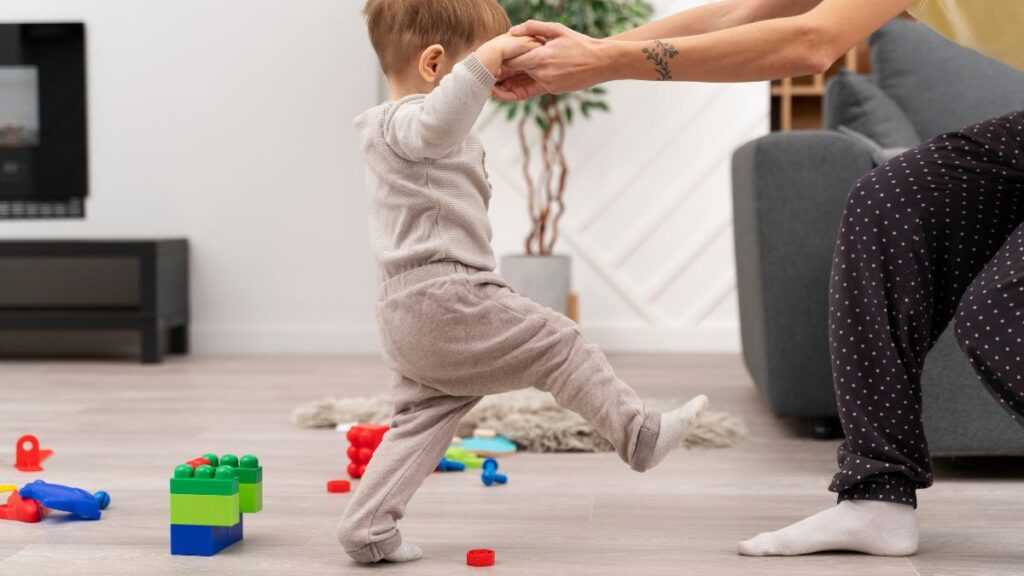 A male toddler walks barefoot on a carpeted floor while a parent gently guides him with both hands. The moment offers a soothing alternative to foot-rubbing behaviors. Colorful toys are scattered nearby.