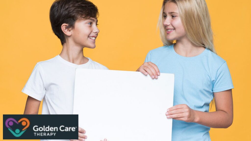Two children, a boy and a girl, stand in a room with a yellow background, holding a blank illustration board together as a visual aid for autism learning activities.