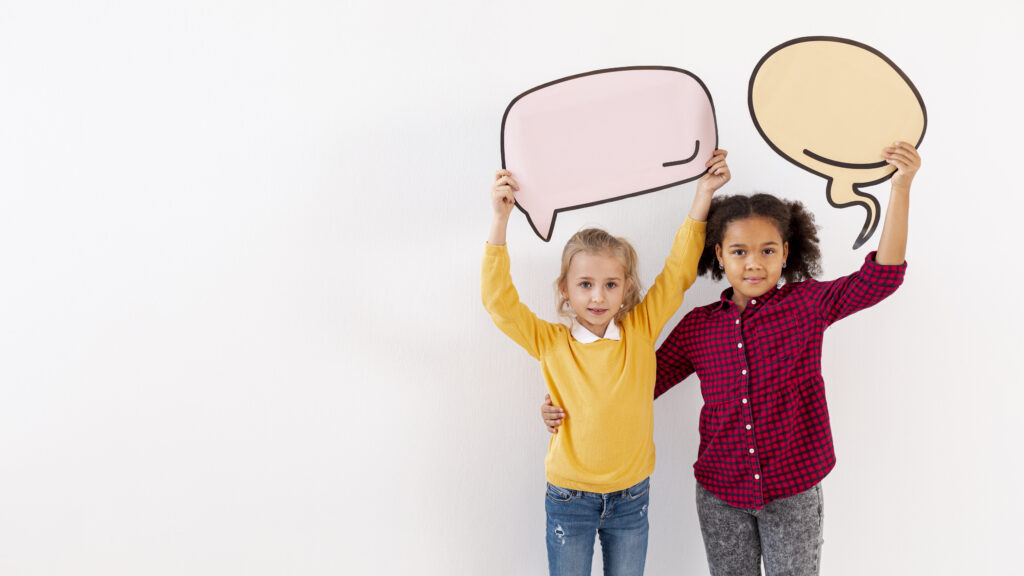 Two young girls with autism hold speech bubbles over their heads while standing close, with hands on each other’s waists, engaging in a language development activity.